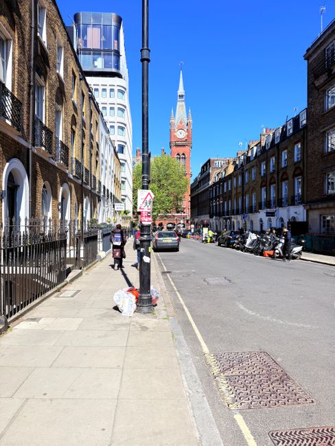 A street scene in West Hampstead with a clear blue sky and a view of a historic red-brick clock tower with a spire, situated in the background. On the left, there are residential buildings with dark iron railings, and on the right, a row of terraced houses with small front gardens and parked vehicles, including motorcycles and cars. The sidewalk is paved with large concrete slabs, and a few pedestrians are visible, one carrying a plastic shopping bag. A black van is parked along the curb, which is adjacent to the pavement. In the foreground, a black pole with street signs and a small traffic warning sign is positioned near the sidewalk. The image captures an urban environment suitable for house removals and furniture transport, with a focus on street accessibility, parking, and loading areas for relocation services offered by Man and Van West Hampstead, in line with Camden Council parking permit regulations.