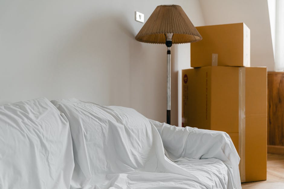 Inside a home during a relocation process, a white fabric cover drapes over a sofa placed against a light-colored wall. To the right of the sofa, there are three cardboard boxes stacked with the larger box at the bottom and smaller ones on top, potentially prepared for transport or packing. Behind the boxes, a tall floor lamp with a brown fabric lampshade is positioned, casting soft light in the room. The scene indicates a typical interior space during moving preparations, with packing materials and furniture arranged for a home relocation. The environment appears neat and organized, with natural light illuminating the scene from a window on the right side. Man and Van West Hampstead, a professional removals service, would manage the furniture transport and packing logistics involved in such a move, ensuring a smooth home relocation experience.