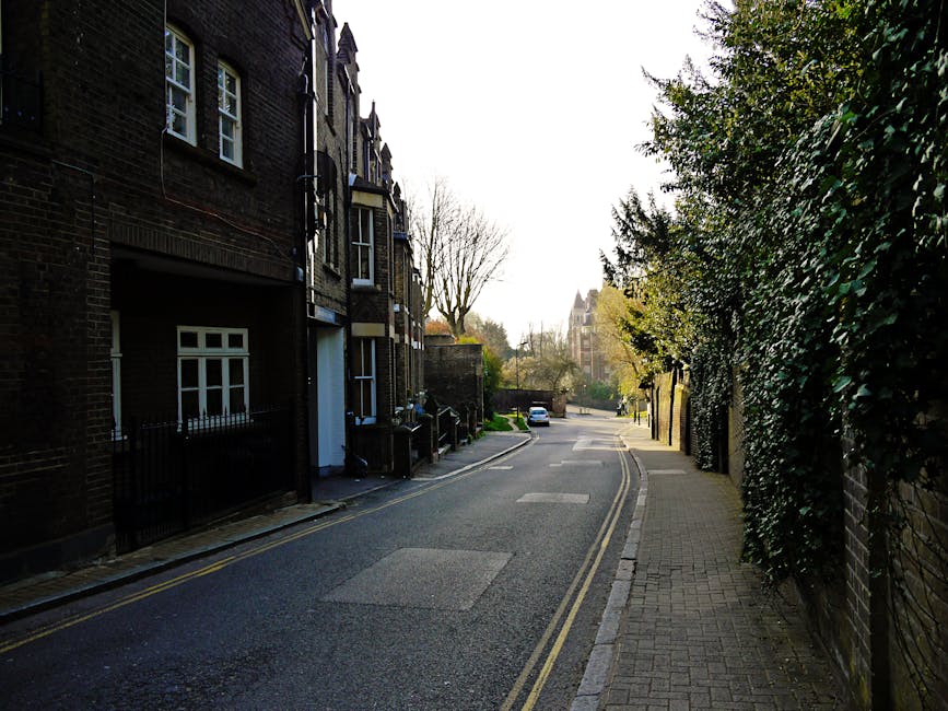 A quiet residential street with narrow pavement on the right and a two-lane road on the left, lined by dark brick terraced houses with white-framed windows and small front gardens, some featuring black metal railings. Tall, leafless trees are visible in the background amidst a clear sky, suggesting early spring or late autumn. The street appears calm, with a few parked cars further along the road. The scene is illuminated by natural daylight, casting soft shadows on the pavement and buildings. This setting is typical of West Hampstead, providing a context for local house removals and furniture transport. The calm and orderly environment reflects the typical conditions encountered during home relocation processes, emphasizing the importance of professional removals services such as those offered by Man and Van West Hampstead, who handle packing, loading, and transportation within such urban residential areas.