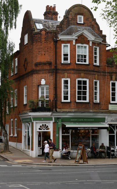 A three-storey red brick building with a curved gable roof and decorative white trim, located on a street corner in West Hampstead. The ground floor features a bakery, café, and pâtisserie with large windows and green signage, with outdoor seating and a chalkboard sign on the sidewalk. On the pavement in front, two people are walking past the shop; one carrying a bag and the other wearing a hat. The building is surrounded by trees, and a van belonging to Man and Van West Hampstead is parked nearby, indicating a home or office relocation process involving furniture transport and packing during a local moving service in the area.
