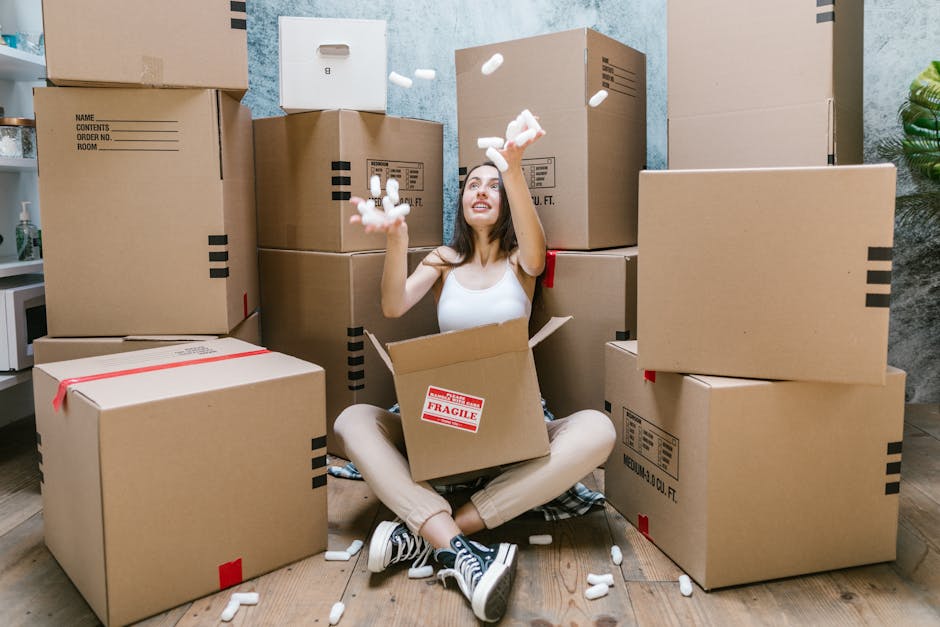 A man and a woman are standing indoors in a room with sloped ceilings, holding large cardboard boxes filled with household items ready for a home relocation. The man, with dark hair and a beard, is we