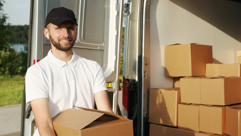 A man wearing a white polo shirt and a black cap is standing outdoors next to an open moving van during daytime, holding a cardboard box in his hands. The van's interior is visible, with several other cardboard boxes stacked inside, some wrapped in plastic film. The scene takes place on a paved driveway with a grassy area and trees in the background, under natural sunlight. The loading process appears to be underway, with the man either preparing boxes for home relocation or furniture transport as part of a professional removals service. Man and Van West Hampstead, a company specializing in removals, is engaged in a home moving or packing and moving operation, utilizing equipment such as trolleys or straps if visible. The focus remains on the loading activity and the organization of packing materials, illustrating the logistics involved in house removals and furniture transport.