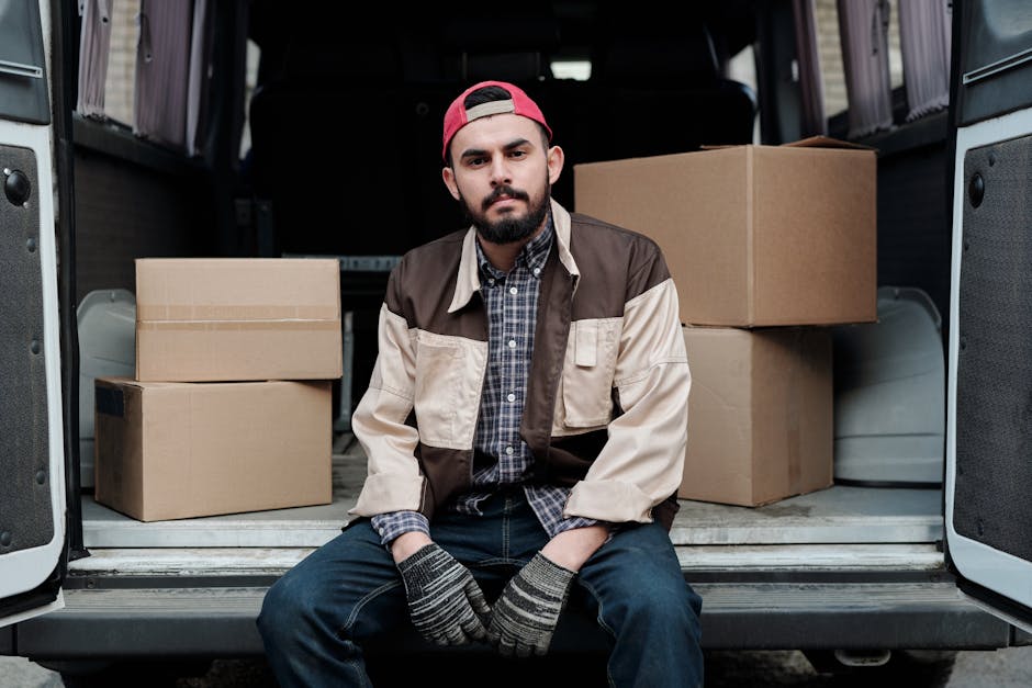 A man with a beard, wearing a red baseball cap backwards, a plaid shirt, and a beige and brown jacket, is seated at the rear of a loading van during a home relocation. The van’s open doors reveal several cardboard boxes of various sizes, some stacked on each other, with packing materials visible inside. The man is sitting on the edge of the vehicle's loading area, facing forward, with his hands in grey striped gloves resting on his knees. The background shows the interior of the van with dark upholstery and some additional unidentified packaging or equipment. The scene is set outdoors, with natural lighting highlighting the moving process involving furniture transport and packing activities, and appears to be part of a professional removals service by Man and Van West Hampstead performing a same-day moving operation.