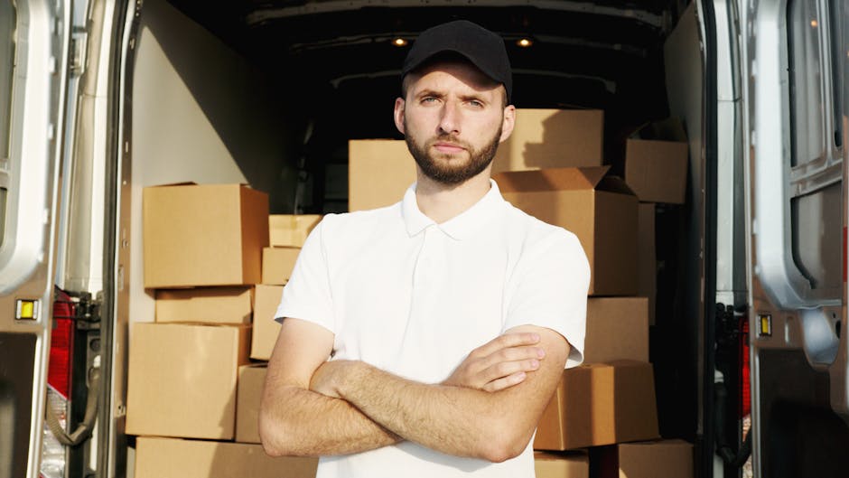 A man wearing a white polo shirt and a black cap is standing outdoors next to an open moving van during daytime, holding a cardboard box in his hands. The van's interior is visible, with several other cardboard boxes stacked inside, some wrapped in plastic film. The scene takes place on a paved driveway with a grassy area and trees in the background, under natural sunlight. The loading process appears to be underway, with the man either preparing boxes for home relocation or furniture transport as part of a professional removals service. Man and Van West Hampstead, a company specializing in removals, is engaged in a home moving or packing and moving operation, utilizing equipment such as trolleys or straps if visible. The focus remains on the loading activity and the organization of packing materials, illustrating the logistics involved in house removals and furniture transport.