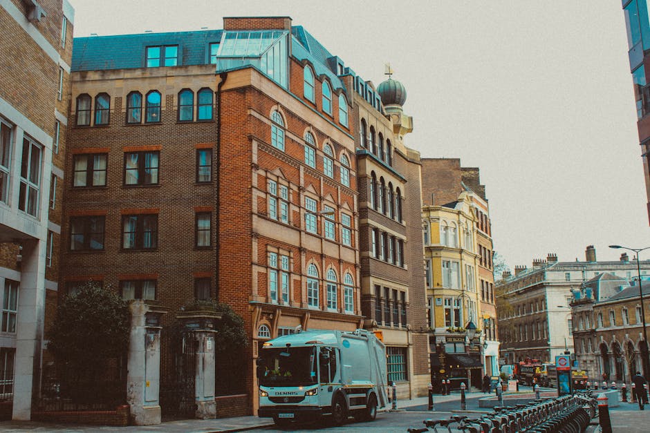 A city street scene showing a grey commercial van parked outside a multi-storey brick building with large windows and a green rooftop. The van is positioned near the entrance of an apartment complex, likely being loaded or unloaded with furniture and boxes for a home relocation. Nearby, there are several other old and ornate buildings with varied architectural styles, including one with a cream and yellow facade and decorative elements. The street is lined with bollards, a row of bicycle racks, and a lamppost, with a few pedestrians and vehicles in the background. The scene is illuminated by daytime natural light, highlighting the urban environment typical of a busy West Hampstead neighbourhood. This image, associated with Man and Van West Hampstead, reflects the logistics involved in furniture transport and packing during removals, emphasizing the importance of careful loading and street-level access in moving services.