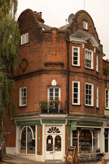 The exterior of a brick residential building with four white-framed windows, two on the upper floor and two on the lower, one of which is partially covered by a sign that reads 'Holly Mount House'. In front of the entrance, there is a set of dark, weathered stone steps with moss and small plants growing in the cracks, leading up to a red wooden door with a small glass pane and a number plaque. To the right of the steps, there is a black metal railing and a small, green rectangular planter with a shrub placed on the paved area beside the door. The surrounding environment includes an old-fashioned street lamp on the left side, and the weather appears damp and overcast, consistent with typical UK conditions suitable for home relocation and furniture transport preparations. This scene is representative of house entryways often seen during relocation or moving processes, with the placement of packing materials and clear access to furniture loading zones nearby, supported by the context of a removal service like Man and Van West Hampstead.