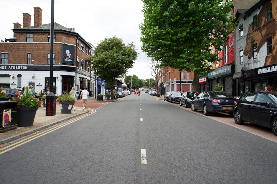 A straight view of a street in West Hampstead with a variety of parked cars lining both sides of the road. On the left side, there are pedestrians walking along the sidewalk, some next to large planters filled with colorful flowers, while a person in a white top and shorts is visible. The buildings on both sides are a mix of commercial establishments and residences, with signs for shops, a pharmacy, and cafes. The street features a central dashed white line, and there are two lush green trees in the middle distance, providing partial shade. The scene appears to be during daytime with clear, natural lighting. This setting exemplifies a typical urban environment involved in local home relocation or furniture transport activities, where [COMPANY_NAME], such as Man and Van West Hampstead, might facilitate moving logistics and packing operations involving loading and unloading at residential or commercial properties.