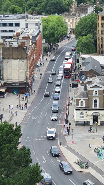 A straight view of a street in West Hampstead with a variety of parked cars lining both sides of the road. On the left side, there are pedestrians walking along the sidewalk, some next to large planters filled with colorful flowers, while a person in a white top and shorts is visible. The buildings on both sides are a mix of commercial establishments and residences, with signs for shops, a pharmacy, and cafes. The street features a central dashed white line, and there are two lush green trees in the middle distance, providing partial shade. The scene appears to be during daytime with clear, natural lighting. This setting exemplifies a typical urban environment involved in local home relocation or furniture transport activities, where [COMPANY_NAME], such as Man and Van West Hampstead, might facilitate moving logistics and packing operations involving loading and unloading at residential or commercial properties.